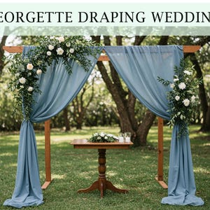 Peut inclure: Une arche de mariage en bois drapée de tissu bleu fluide et ornée de fleurs blanches et de verdure. Une petite table en bois avec des compositions florales et des bougies se trouve sous l'arche. Le texte "GEORGETTE DRAPING WEDDING" est en haut.