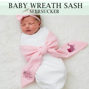 Könnte beinhalten: Ein Neugeborenes in einer weißen Windel mit einer rosa-weiß gestreiften Seersucker-Schärpe. Die Schärpe ist zu einer Schleife gebunden und zeigt einen gestickten Kinderwagen und den Text "Little Princess". Das Baby trägt ein passendes Stirnband. Der Text "BABY WREATH SASH SEERSUCKER" steht oben.