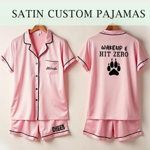 Op de afbeelding: Roze satijnen pyjama met zwarte bies. Het bovenstuk heeft een knoopsluiting en een zakje met de naam "Michelle" geborduurd erop. De achterkant van het bovenstuk heeft de tekst "Wake Up & Hit Zero" en een zwarte pootprint. De broek heeft het woord "Cheer" op de achterkant gedrukt.