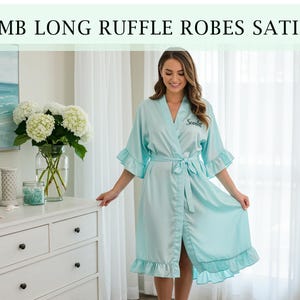 Pode incluir: Robe de cetim azul claro com babados nas mangas e na bainha. O robe tem um cinto a condizer e a palavra "Scarlett" bordada no peito. O texto "EMB LONG RUFFLE ROBES SATIN" est&aacute; na parte superior da imagem.