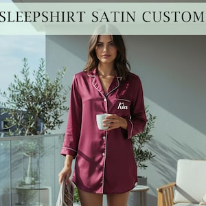 May include: Burgundy satin sleepshirt with white trim and the name "Kia" embroidered on the chest. The sleepshirt is button-down and has long sleeves. The text "SLEEPSHIRT SATIN CUSTOM" is at the top of the image.