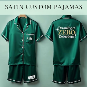 May include: Green satin pajamas with white piping. The top has a pocket with the name "Lily" embroidered on it. The back of the top has the text "Dreaming of ZERO Deductions" in gold lettering. The shorts have the word "CHEER" printed on the leg.