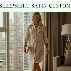 Peut inclure: Une chemise de nuit en satin crème avec une bordure noire et le texte "SLEEPSHIRT SATIN CUSTOM". La chemise de nuit a une fermeture boutonnée et un nom personnalisé sur la poche. Le modèle tient un livre.