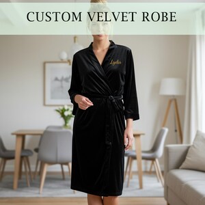 May include: A black velvet robe with a wrap-around design and a matching belt. The robe has long sleeves and a gold embroidered name on the chest. The text "CUSTOM VELVET ROBE" is displayed at the top of the image.