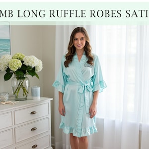 Pode incluir: Um roup&atilde;o de cetim azul claro com mangas e bainha com babados. O roup&atilde;o tem um cinto a condizer e o texto "Sanka" bordado no peito. A imagem tamb&eacute;m inclui o texto "EMB LONG RUFFLE ROBES SATIN".