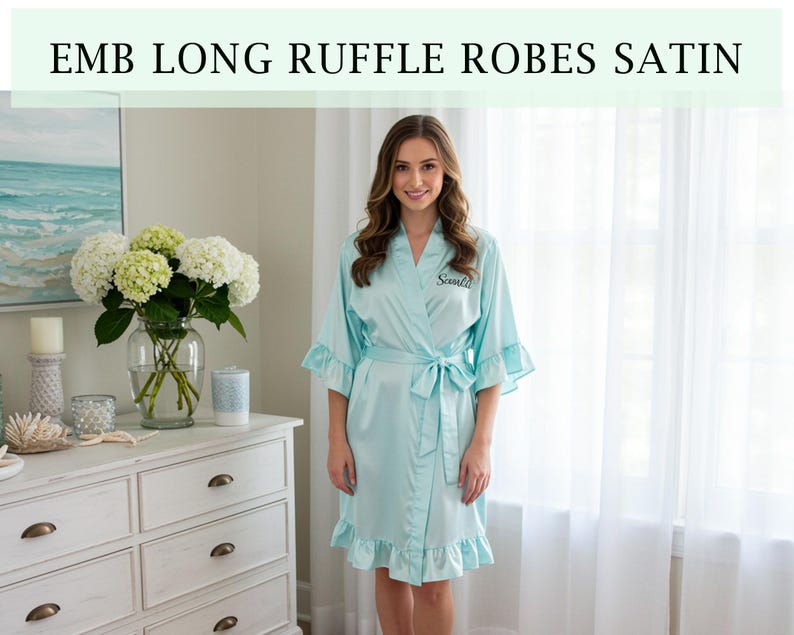 Pode incluir: Um roup&atilde;o de cetim azul claro com mangas e bainha com babados. O roup&atilde;o tem um cinto a condizer e o texto "Sanka" bordado no peito. A imagem tamb&eacute;m inclui o texto "EMB LONG RUFFLE ROBES SATIN".