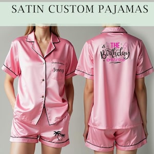 May include: Pink satin pyjamas with black trim. The top has a button fastening and a pocket with the name "Jenny" embroidered on it. The back of the top has the text "The Birthday Squad" in black and pink lettering. The shorts have a palm tree design.