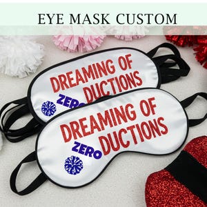 May include: Two white satin eye masks with black elastic straps. The masks have the text "Dreaming of Ductions" in red glitter lettering and a blue and white cheerleading pom-pom graphic. The text "Zero" is also included in the graphic.