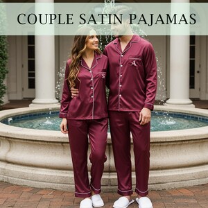 Könnte beinhalten: Passende bordeauxfarbene Satin-Pyjamas mit weißem Paspel. Die langärmligen Hemden mit Knöpfen und Hosen werden von einem Paar getragen. Der Text "COUPLE SATIN PAJAMAS" befindet sich oben im Bild. Jedes Pyjama-Oberteil hat eine einzelne Initiale.