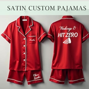 May include: Red satin pajamas with white piping. The top has a pocket with the name "Kendra" embroidered on it. The back of the top has the text "Wakeup & HIT ZERO" with a white megaphone graphic. The shorts have the text "Cheer Leader" embroidered on them.
