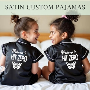 Peut inclure: Deux enfants portant des pyjamas en satin noir avec des bordures blanches et un motif de papillon. Les pyjamas ont le texte "Wake up & HIT ZERO" au dos.