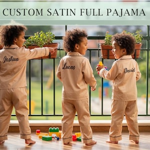 Könnte beinhalten: Drei Kinder tragen passende, hellbraune Satin-Schlafanzüge mit schwarzem Besatz und personalisierten Namen auf dem Rücken. Die Schlafanzüge sind mit "Joshua", "Lucas" und "David" beschriftet. Das Bild enthält den Text "CUSTOM SATIN FULL PAJAMA".