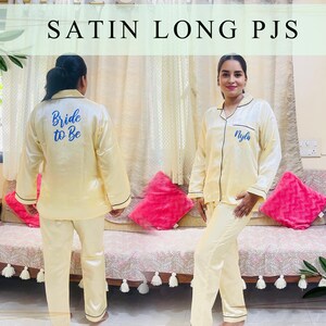 May include: Two people wearing matching long-sleeved satin pyjamas. One pyjama top has the text "Bride to Be" embroidered on the back in blue thread. The other pyjama top has the name "Nyla" embroidered on the front in blue thread.