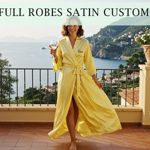 May include: A woman wearing a yellow satin robe with the name "Kia" embroidered on the front. The robe is open, revealing the legs. The text "FULL ROBES SATIN CUSTOM" is at the top of the image. The background shows a scenic view of the ocean and mountains.
