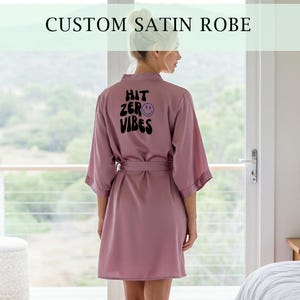 May include: A mauve satin robe with a matching belt, featuring the text "HIT ZERO VIBES" on the back. The robe has wide sleeves and is knee-length. The top of the image reads "CUSTOM SATIN ROBE".