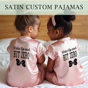 Peut inclure: Deux filles portant des pyjamas en satin rose avec le texte "Wake Up and HIT ZERO" et un nœud noir au dos.