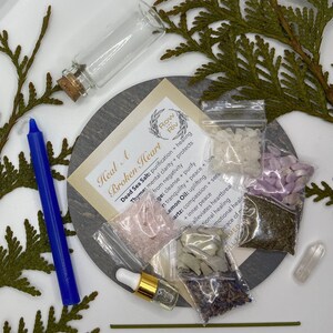 Puede incluir: Un conjunto de cristales curativos, hierbas y aceites esenciales para un ritual de sanación del corazón roto. El conjunto incluye una pequeña botella de aceite esencial transparente, una pequeña bolsa de sal rosa del Himalaya, una pequeña bolsa de cristales blancos, una pequeña bolsa de capullos de lavanda, una pequeña bolsa de hierbas verdes y una pequeña botella de vidrio transparente con un tapón de corcho. El conjunto está dispuesto sobre una superficie circular gris con las palabras "Heal A Broken Heart" escritas en una tarjeta blanca.