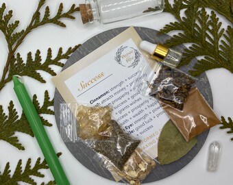 May include: A success spell kit with a green candle, a small glass bottle, a clear quartz crystal, a bay leaf, and four small bags containing herbs and spices. The bags are labeled with the words "Cinnamon", "Eye", "Oil", and "Incense". The kit also includes a small card with the word "Success" written on it.