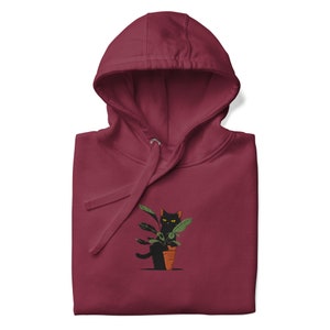 Könnte beinhalten: Ein weinroter Hoodie mit einer schwarzen Katze, die aus einer Topfpflanze schaut. Die Katze hat gelbe Augen und die Pflanze hat grüne Blätter.