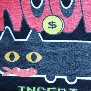 May include: Black t-shirt with a cartoon cat design. The cat has yellow eyes and a red mouth. Above the cat is a yellow coin with a dollar sign. The word "WOW" is in red above the cat.