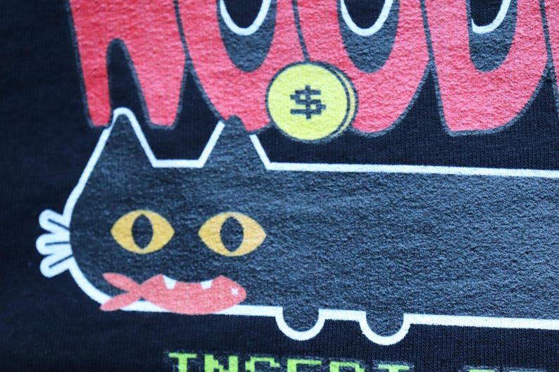 May include: Black t-shirt with a cartoon cat design. The cat has yellow eyes and a red mouth. Above the cat is a yellow coin with a dollar sign. The word "WOW" is in red above the cat.