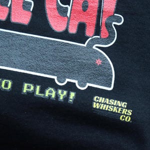 May include: Black t-shirt with a retro-style graphic. The design features the words "OLDER CAT" in red, a gray cat-shaped design, and the text "5&cent; TO PLAY!" in green. The shirt also includes the text "CHASING WHISKERS CO."