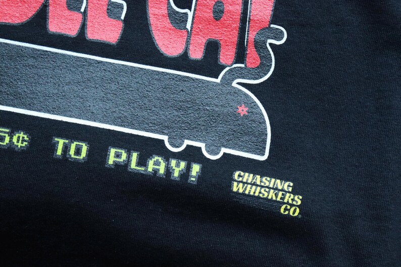 May include: Black t-shirt with a retro-style graphic. The design features the words "OLDER CAT" in red, a gray cat-shaped design, and the text "5&cent; TO PLAY!" in green. The shirt also includes the text "CHASING WHISKERS CO."