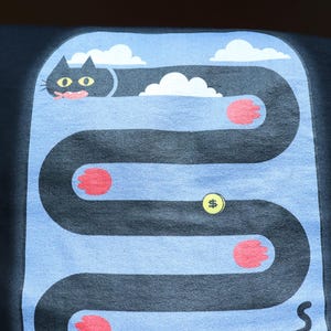 May include: Black t-shirt with a whimsical cat graphic. The design features a black cat with yellow eyes, a pink tongue, and red paws, navigating a winding path with clouds and a dollar sign.