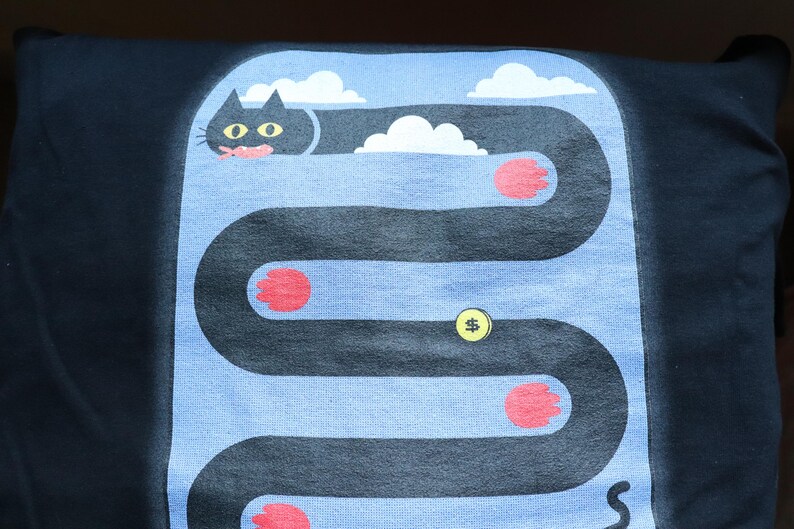 May include: Black t-shirt with a whimsical cat graphic. The design features a black cat with yellow eyes, a pink tongue, and red paws, navigating a winding path with clouds and a dollar sign.
