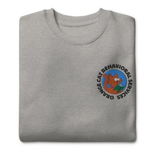 May include: A grey crew neck sweatshirt. It has an embroidered circular patch with the text "ORANGE CAT BEHAVIORAL SERVICES" and a cartoon orange cat reading a book. The sweatshirt is a casual style.