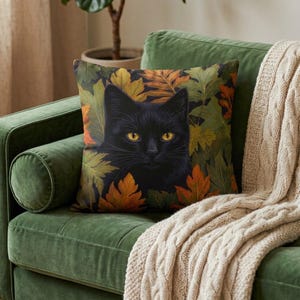 Pode incluir: Uma almofada quadrada com uma ilustração detalhada de um gato preto com olhos amarelo-brilhantes. O gato está rodeado por um padrão denso de folhas de outono em tons de laranja, amarelo e verde, sobre um fundo escuro.