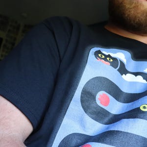 May include: Navy blue t-shirt featuring a cartoon snake design with a cat's head, red accents, and a gold coin. The snake winds through a light blue background. The shirt is made of a soft material.