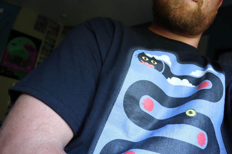 May include: Navy blue t-shirt featuring a cartoon snake design with a cat's head, red accents, and a gold coin. The snake winds through a light blue background. The shirt is made of a soft material.