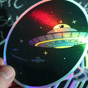 Puede incluir: Pegatina holográfica que representa un OVNI de dibujos animados con un gato dentro, sobre un fondo espacial estrellado. La pegatina tiene un brillo de arcoíris e incluye OVNIs más pequeños en vuelo. El diseño es caprichoso y colorido.