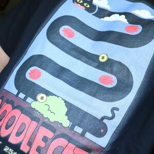 May include: Black t-shirt with a colorful graphic design. The design features a black cat on a winding path with red paw prints, dollar coins, and the text "NOODLE CAT". The shirt also has the text "INSERT 25&cent;".