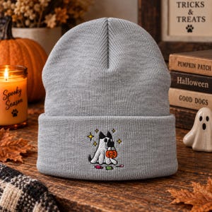 May include: A light gray knit beanie featuring an embroidered graphic of a dog wearing a white ghost costume and holding a small orange pumpkin bucket, surrounded by yellow star accents and scattered colorful candy pieces on the cuff.