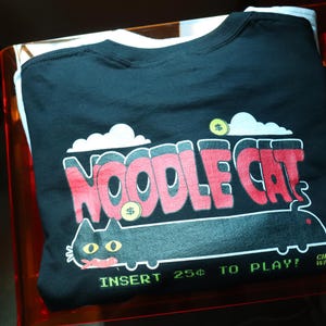 May include: Black t-shirt with a graphic that says "NOODLE CAT" in red and white, with a cartoon cat illustration. The shirt includes the text "INSERT 25&cent; TO PLAY!" and is folded inside a clear orange container.