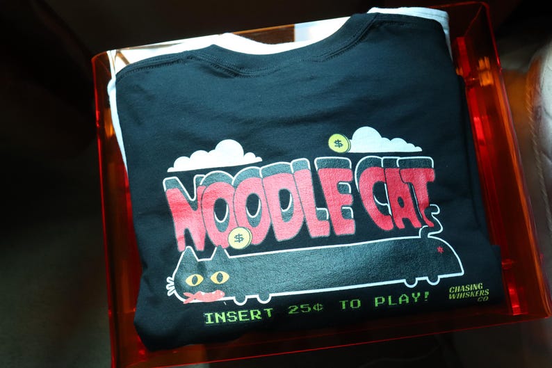 May include: Black t-shirt with a graphic that says "NOODLE CAT" in red and white, with a cartoon cat illustration. The shirt includes the text "INSERT 25&cent; TO PLAY!" and is folded inside a clear orange container.