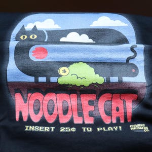 May include: Black t-shirt with a graphic of a long black cat with yellow eyes, a red circle, and a green cloud with a dollar sign. The text "NOODLE CAT" is in red, and "INSERT 25&cent; TO PLAY!" is below.