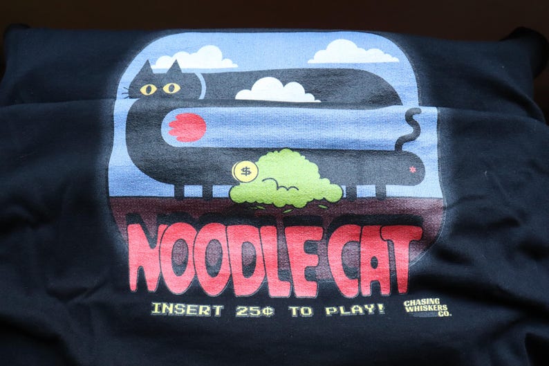 May include: Black t-shirt with a graphic of a long black cat with yellow eyes, a red circle, and a green cloud with a dollar sign. The text "NOODLE CAT" is in red, and "INSERT 25&cent; TO PLAY!" is below.