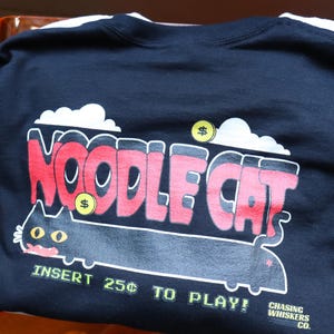 May include: Black t-shirt with a playful "Noodle Cat" graphic in red and white, featuring a cartoon cat and the text "Insert 25&cent; to Play!" The design includes dollar signs and cloud accents. From Chasing Whiskers Co.