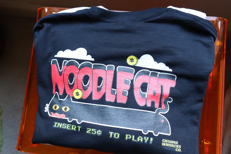 May include: Black t-shirt with a playful "Noodle Cat" graphic in red and white, featuring a cartoon cat and the text "Insert 25&cent; to Play!" The design includes dollar signs and cloud accents. From Chasing Whiskers Co.