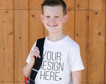 May include: A young person wearing a white t-shirt with the text "YOUR DESIGN HERE" printed on it. They are wearing blue jeans and brown sneakers. They are standing in front of a wooden wall.