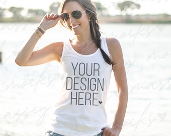 Pode incluir: Um top branco de alças com o texto "YOUR DESIGN HERE" impresso na frente. O top está sendo usado por uma pessoa em pé perto de um corpo de água.