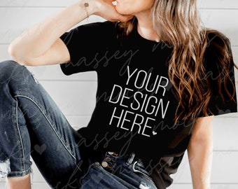 以下が含まれることがあります： 白い文字で「YOUR DESIGN HERE」と書かれた黒いTシャツ。長い茶色の髪の女性が着用しており、白い木製の表面に座っています。