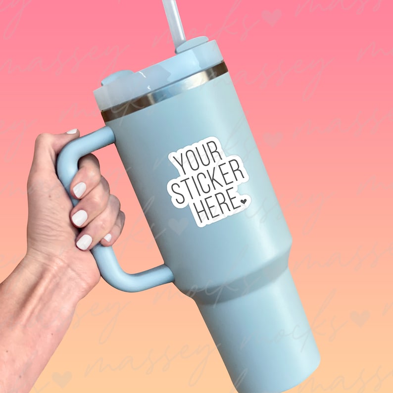 May include: A light blue stainless steel travel mug with a handle and a straw. The mug has a sticker that says "YOUR STICKER HERE" with a small heart.