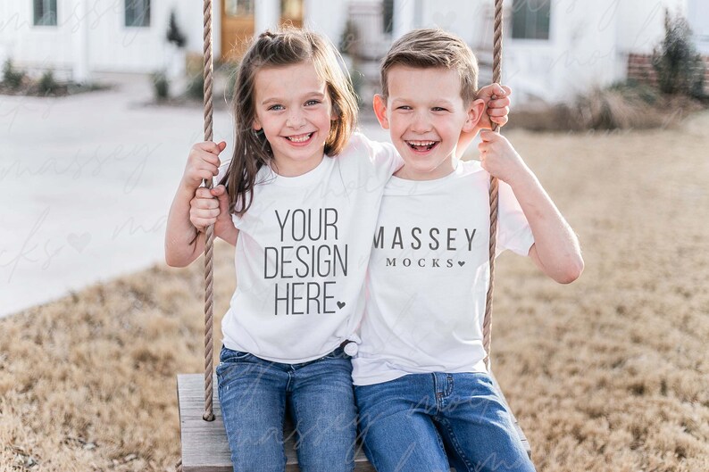 May include: Two children wearing white t-shirts with text on them are sitting on a swing. The girl's shirt says "YOUR DESIGN HERE" and the boy's shirt says "MASSEY MOCKS". They are both smiling and looking at the camera.