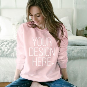 May include: A light pink sweatshirt with the text "YOUR DESIGN HERE" in white. The sweatshirt is a crew neck style and is being worn by a person. The person is wearing blue jeans.