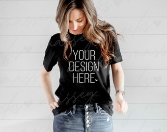 以下が含まれることがあります： 白い文字で「YOUR DESIGN HERE」と書かれた黒いTシャツ。長い茶色の髪をした人が着用しており、青いジーンズと合わせています。