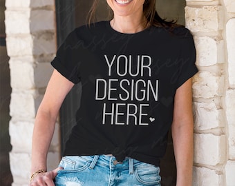 以下が含まれることがあります： 白いハートのマークが付いた白い文字で「YOUR DESIGN HERE」と書かれた黒いTシャツ。長い茶色の髪の女性が着用しており、ウエストで結ばれています。